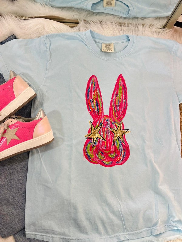 A light blue colored t-shirt with a graphic print of an eye-catching, colorful Easter bunny on it, displayed flat with a pair of pink sneakers and a white shoe on top of it.