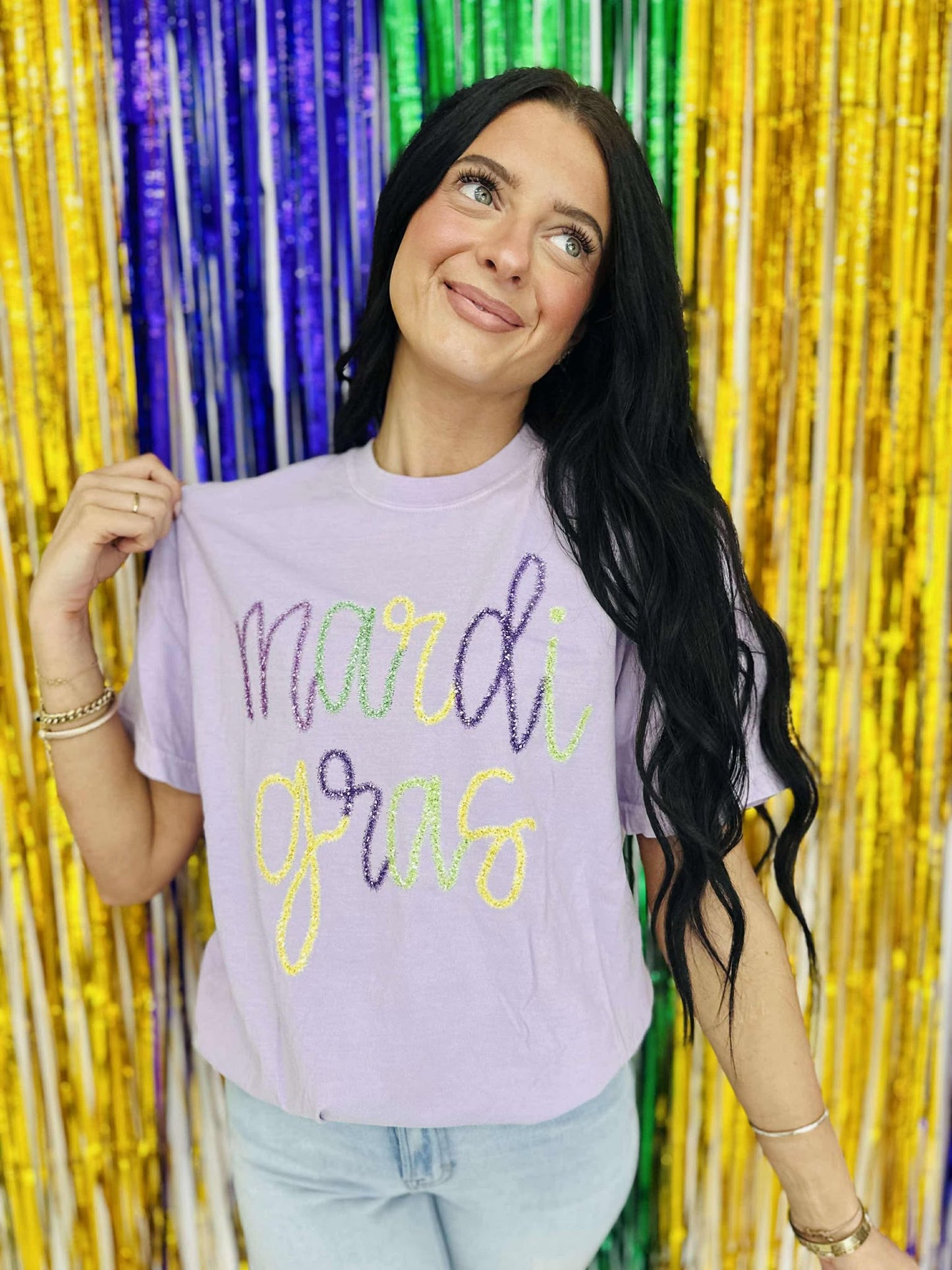 A person holding up a purple t-shirt with 'mardi gras' printed in yellow and neon green tinsel text.