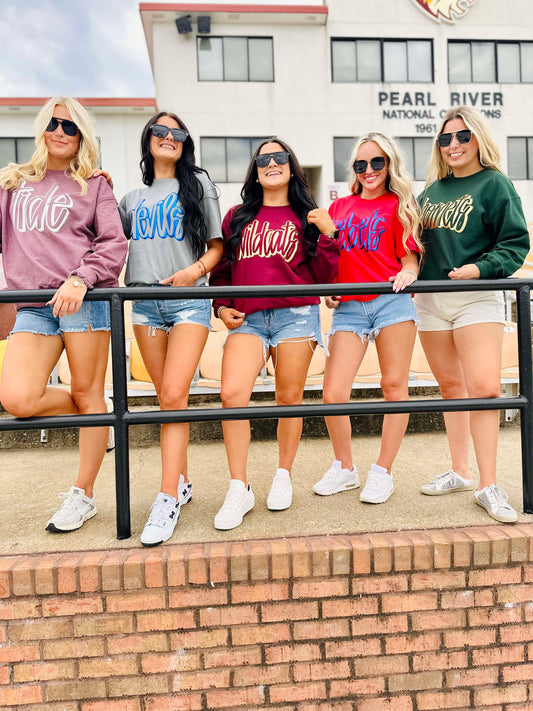 Five women posing together wearing matching sweatshirts in front of a building.