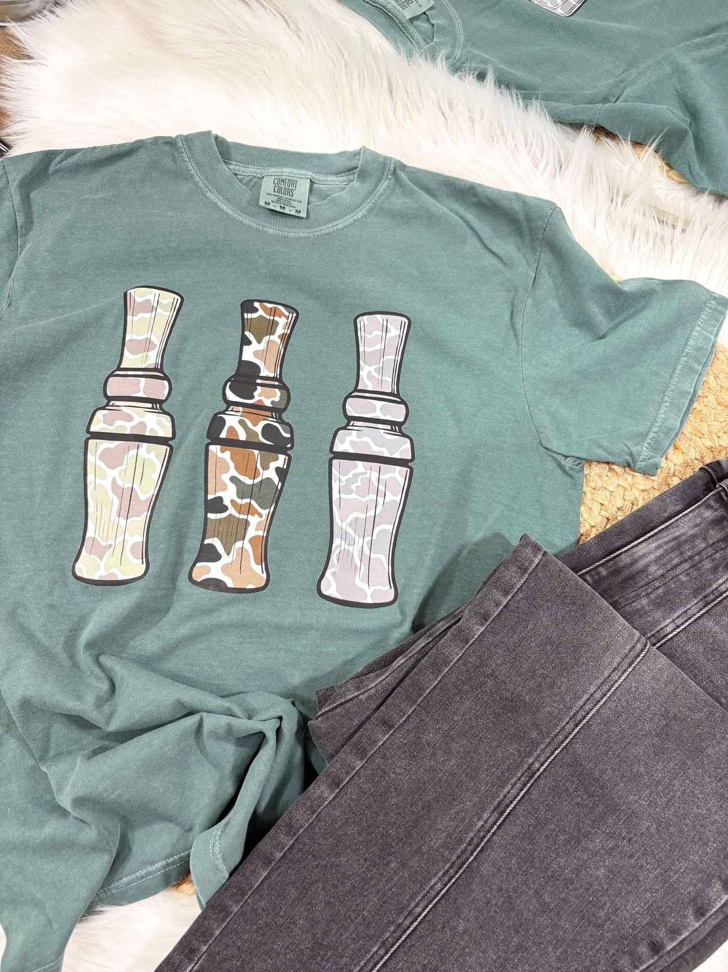Green t-shirt with three camouflaged duck call designs on a white surface.