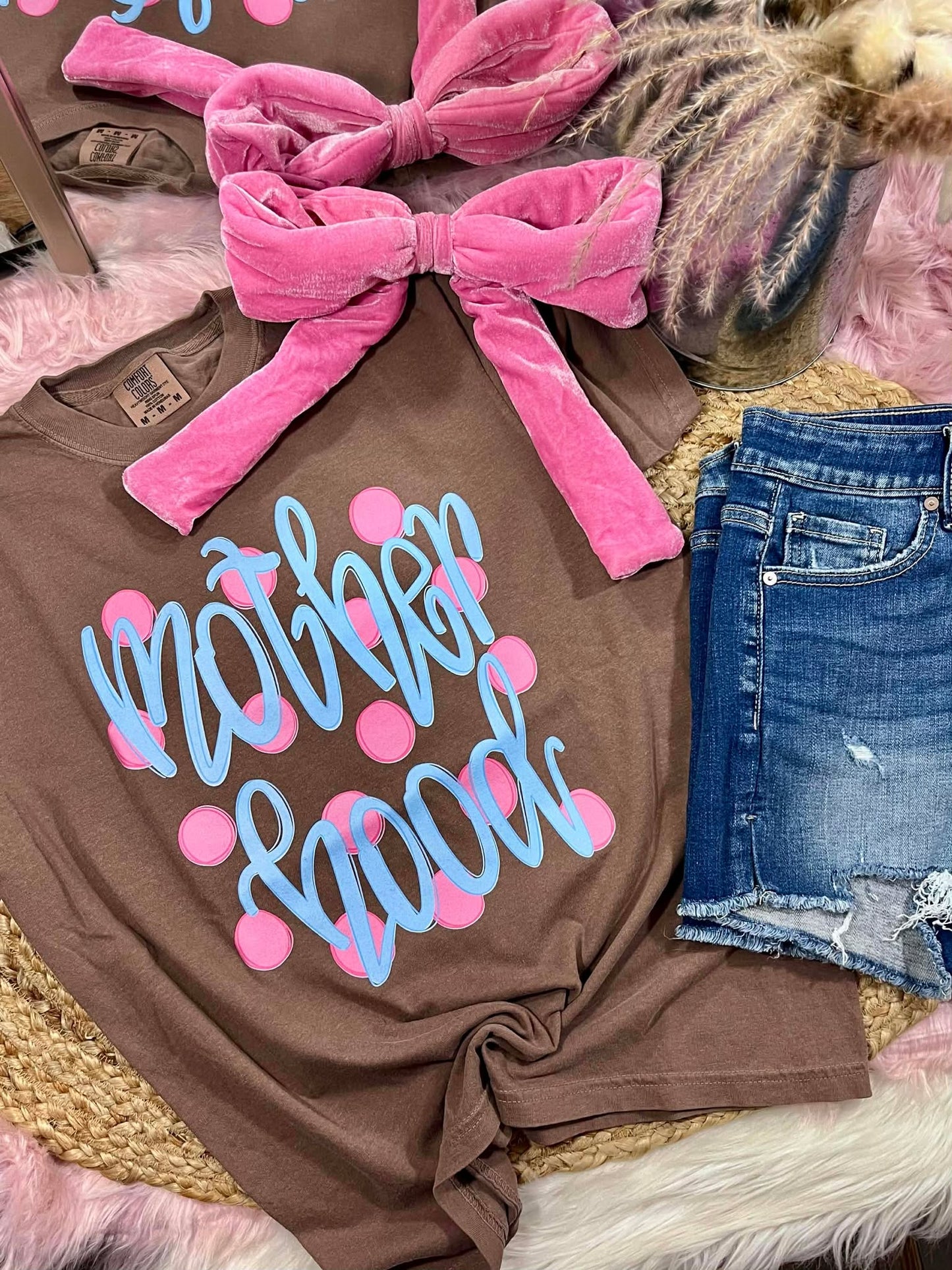 Brown t-shirt with 'Mother Hood' text and pink polka dots, paired with denim shorts and a pink bow on a textured surface.