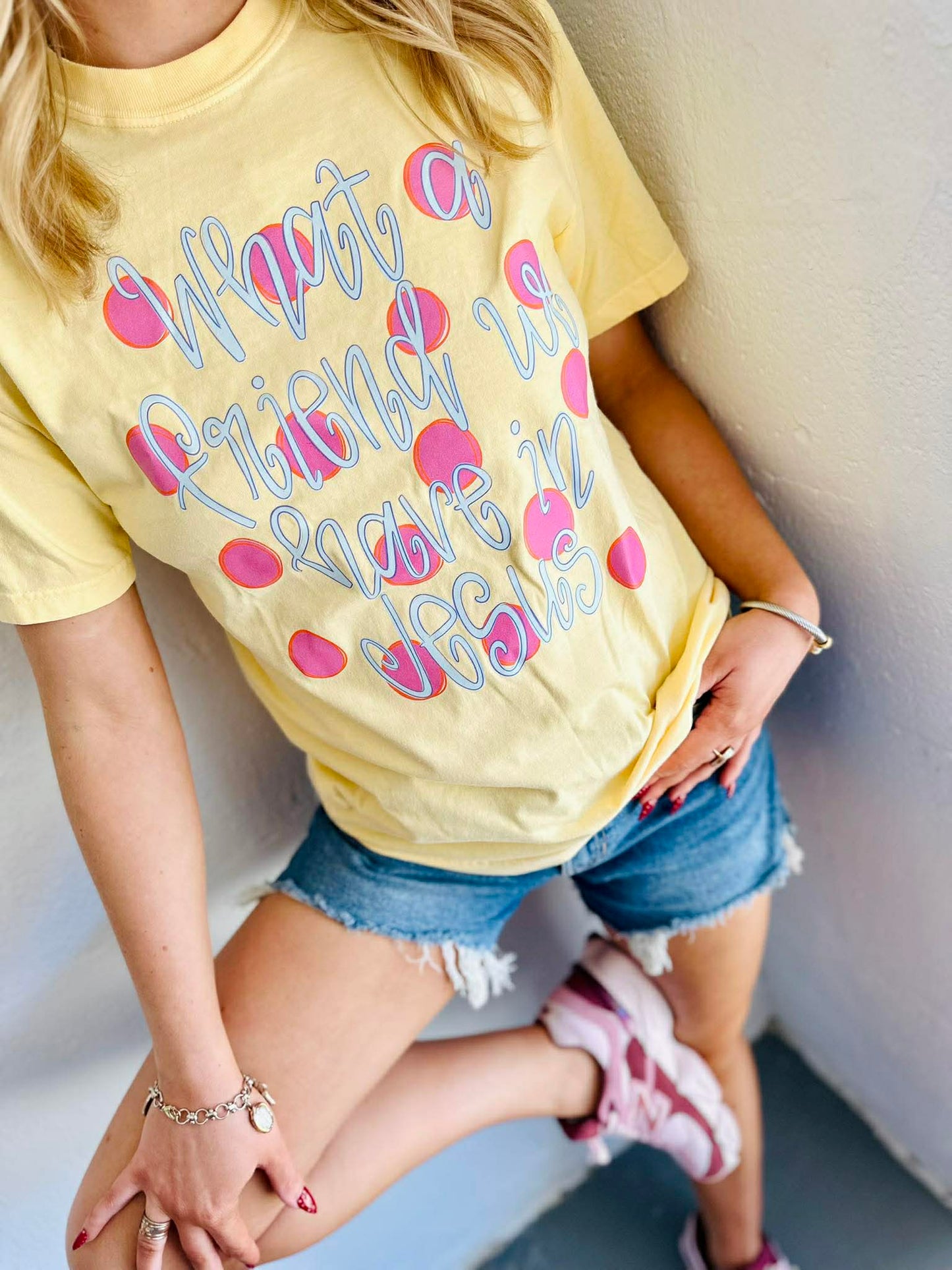 Person wearing a yellow t-shirt with text "What a friend we have in Jesus" polka dots and pink hearts, sitting against a light-colored wall.