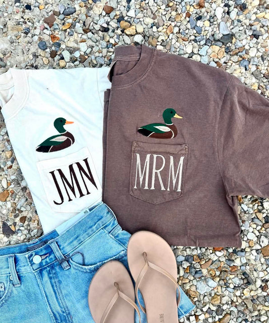 A styled flat-lay of two graphic t-shirts with duck illustrations and pocket monograms, paired with light blue denim shorts and beige flip-flops on a bed of small gray pebbles. One white t-shirt has a mallard graphic above the text 'JMN', and one brown t-shirt has a mallard graphic above the text 'MRM'.