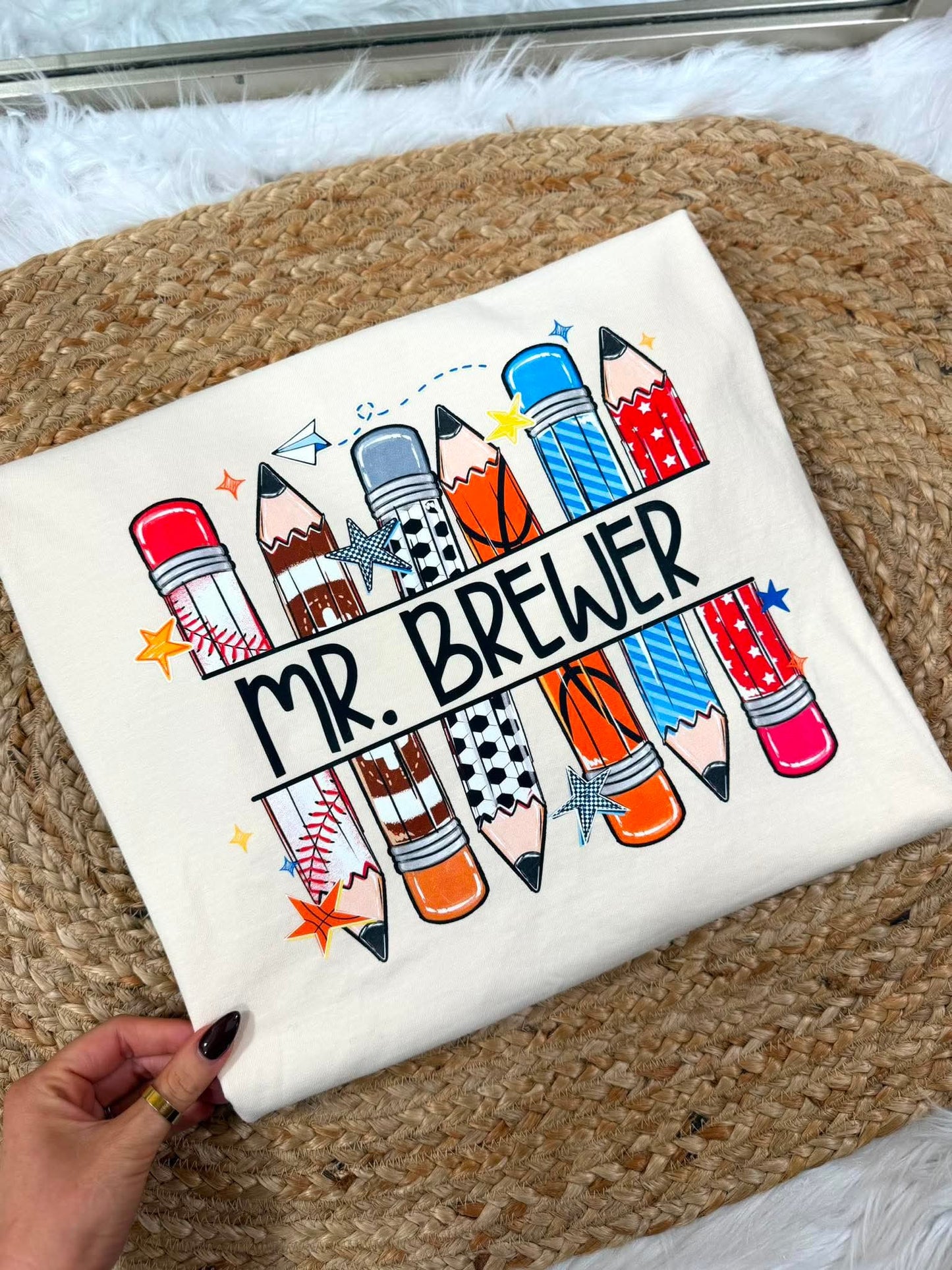 A folded cream-colored t-shirt with a colorful graphic of various illustrated pencils and the text 'MR. BREWER' across the front.