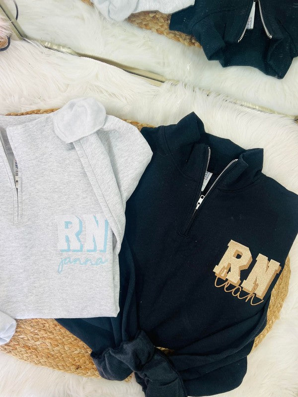 Two hooded jackets, one black and one gray, laid out on a white surface with 'RN' embroidered in gold on the front. Both jackets have a crew neck and a 1/4 zip design.