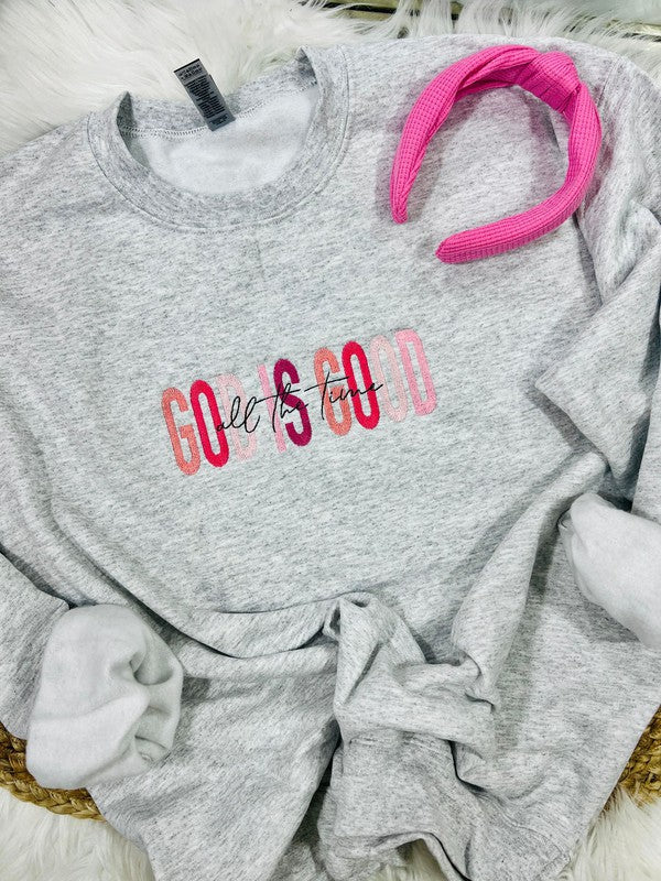 A gray sweatshirt with the phrase 'God Is Good' embroidered in pink on the front, displayed on a white fluffy surface with a pink hairband beside it.