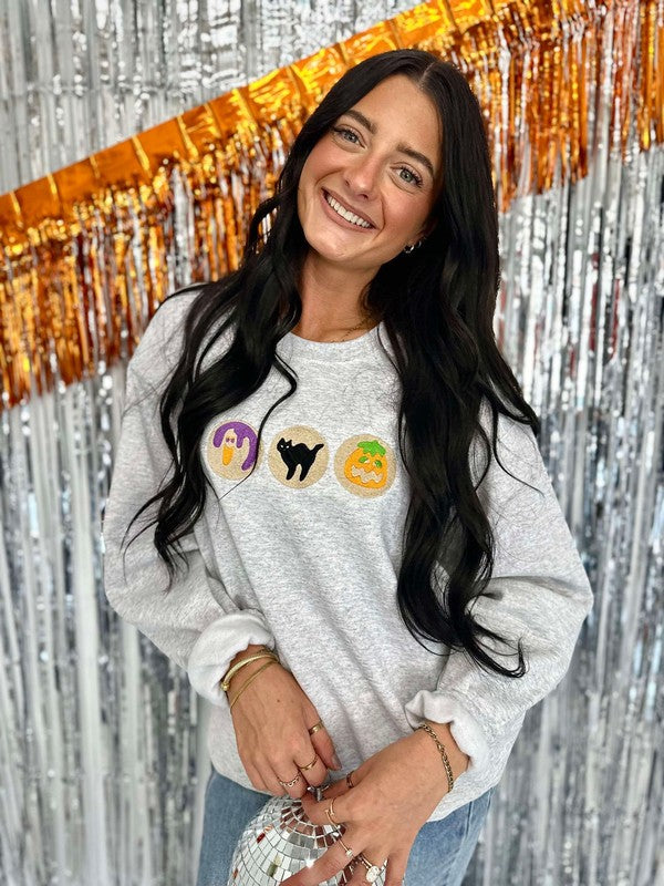 A woman smiling, wearing a long-sleeved gray sweatshirt with orange and black embroidered halloween cookies, and a sparkly background.