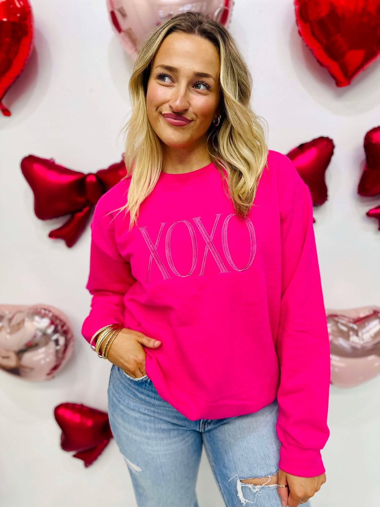 A woman wearing a hot pink embroidered sweatshirt with the text 'XOXO' on it, posing against a background of red and white balloons and decorations.
