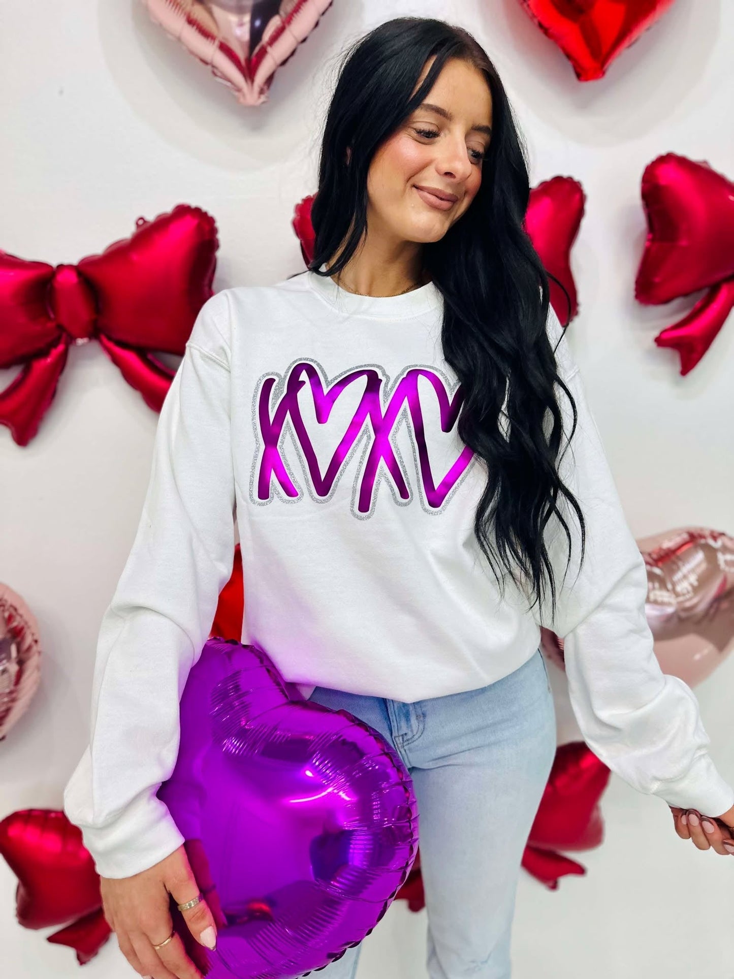 Woman wearing a white sweatshirt with purple text, holding a purple heart-shaped balloon, standing against a wall with red heart decorations.