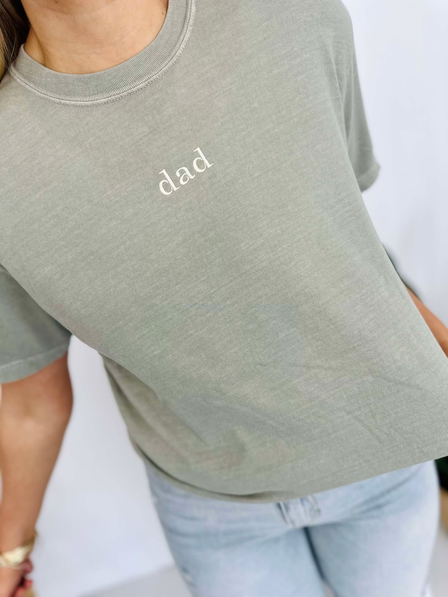 A person wearing a sandstone-colored t-shirt with the word 'DAD' embroidered on the front.