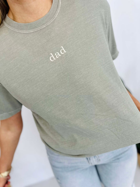 A person wearing a sandstone-colored t-shirt with the word 'DAD' embroidered on the front.