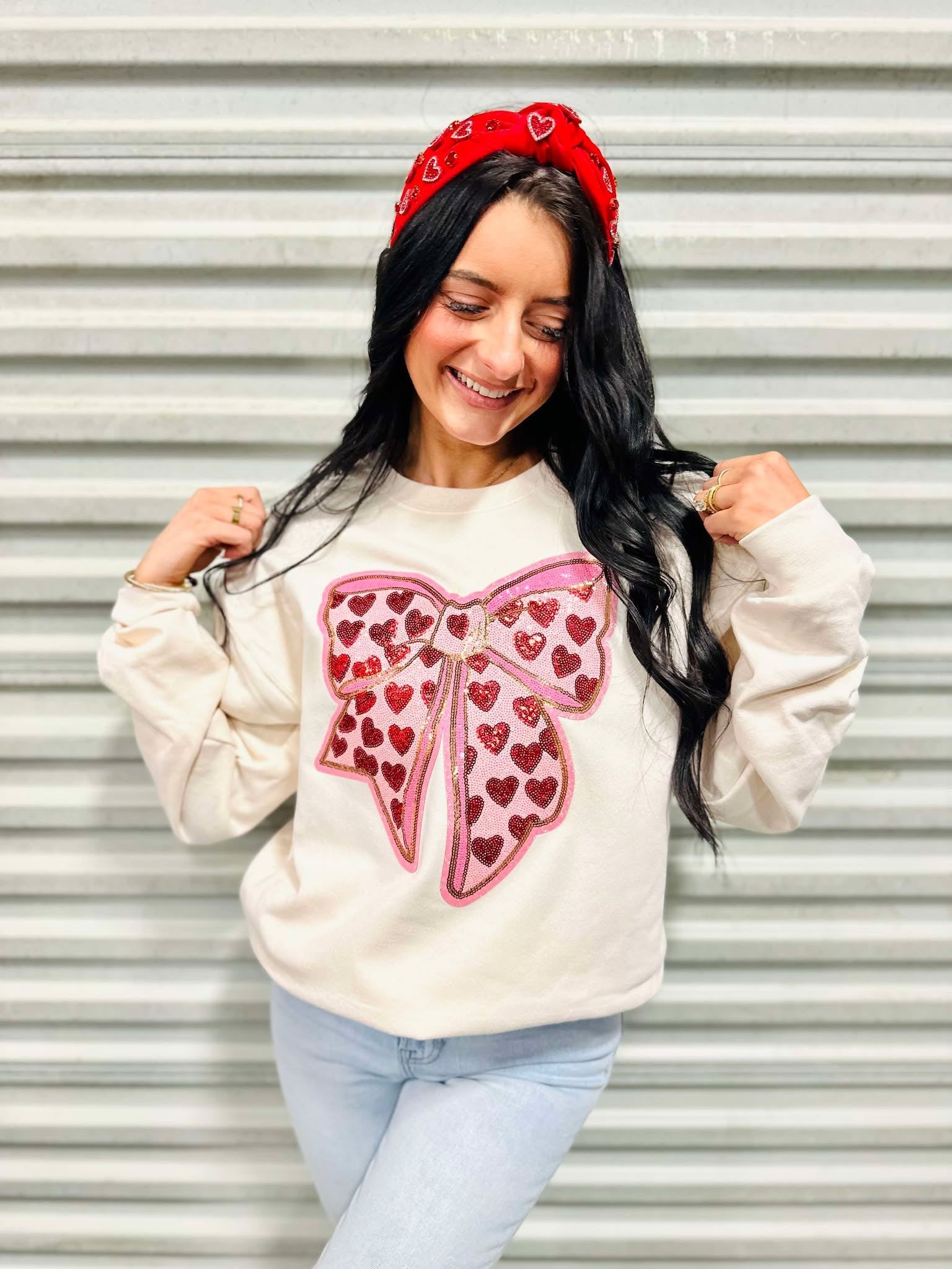 A woman smiling, wearing a beige sweatshirt with a red bow and heart patches, and a red headband.