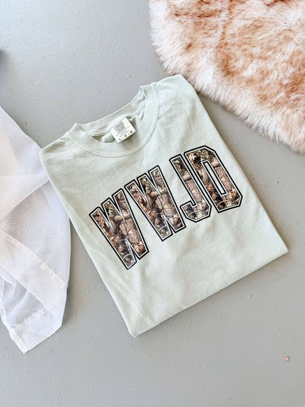 A white t-shirt with 'WWJD' camouflage pattern printed on it, displayed flat with a tag and a furry item in the background.