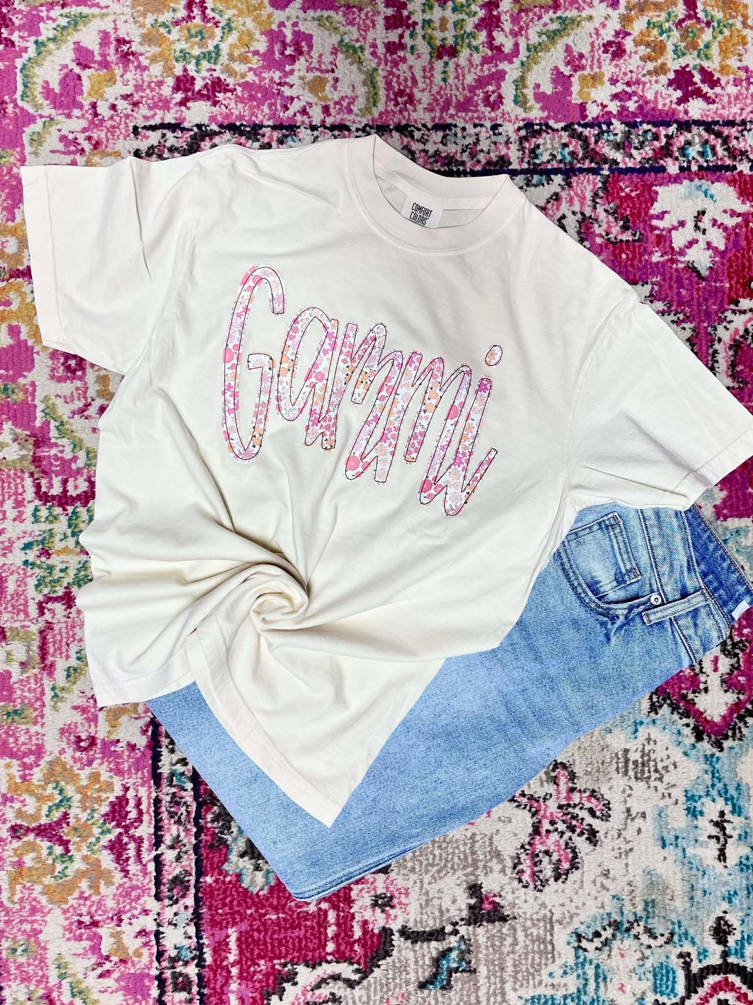 White t-shirt with colorful 'Gammi' text on a multicolored patterned surface