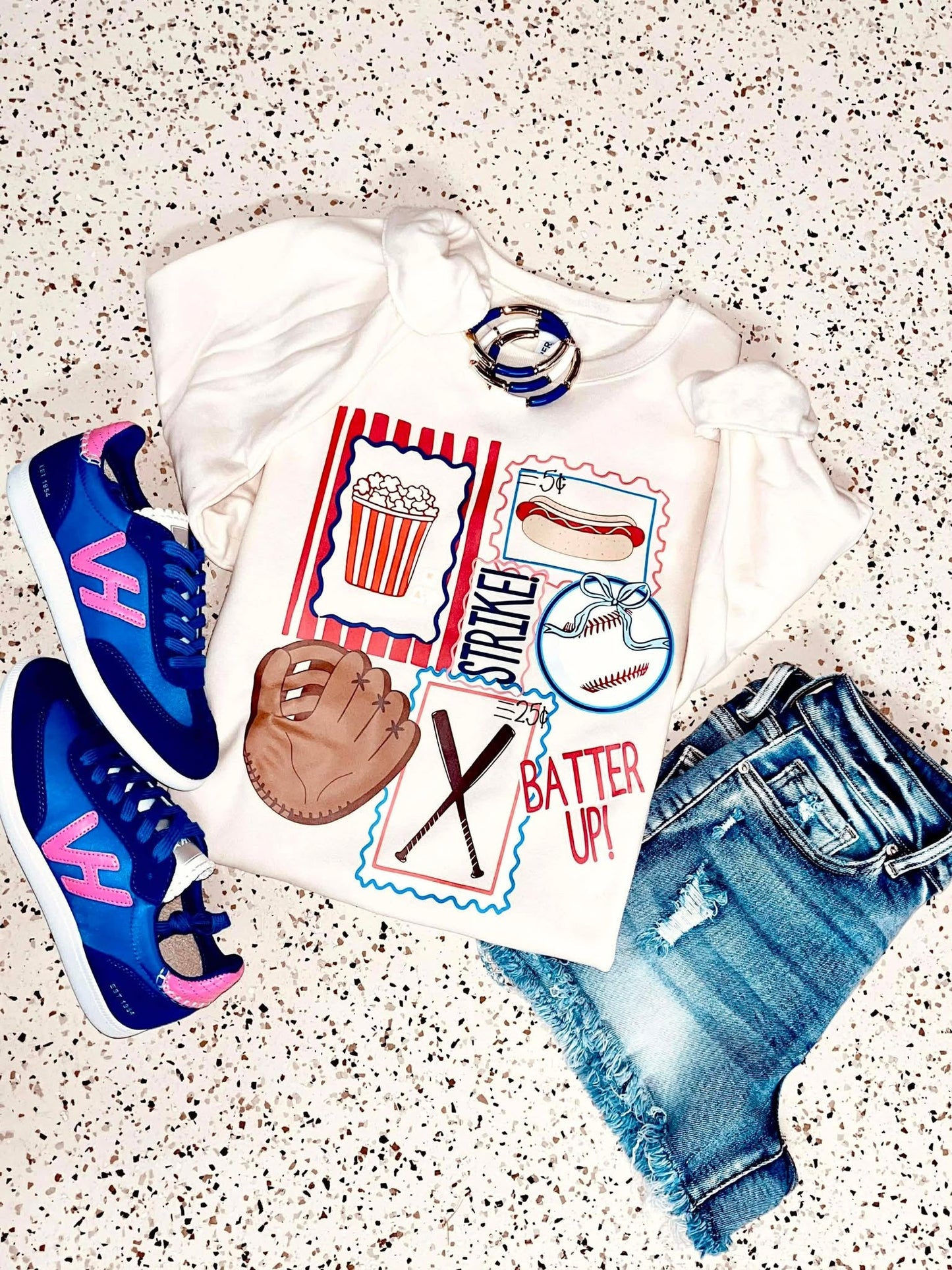 White t-shirt with sports-themed design, blue sneakers, baseball glove, and denim shorts on a speckled surface.