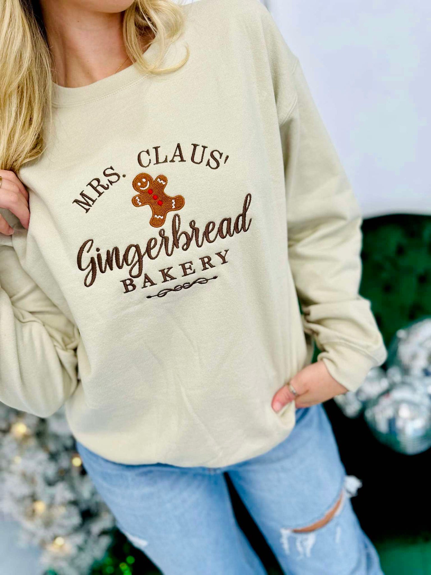 A woman wearing a beige embroidered sweatshirt with 'MRS. CLAUS' GINGERBREAD BAKERY' text and a cute gingerbread man graphic.