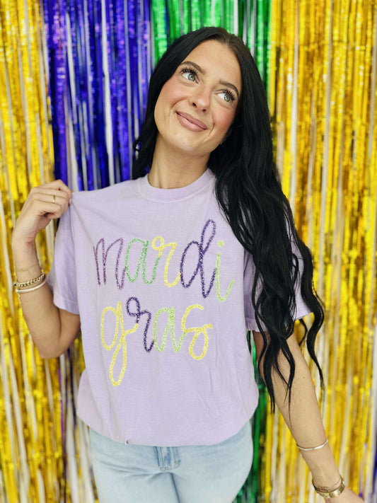 A person holding up a purple t-shirt with 'mardi gras' printed in yellow and neon green tinsel text.