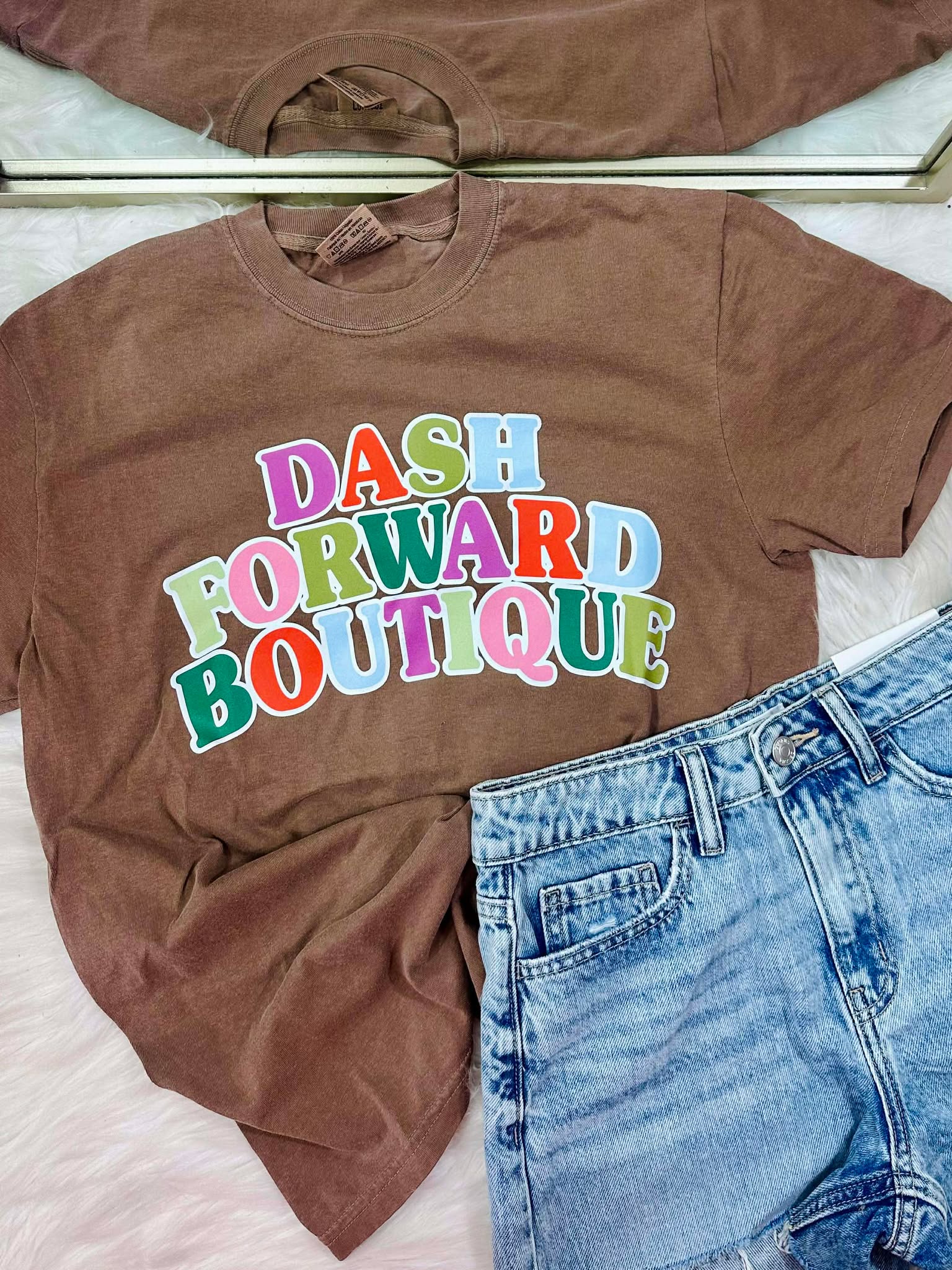 Brown t-shirt with 'Dash Forward Boutique' text and blue jeans on a light pink surface