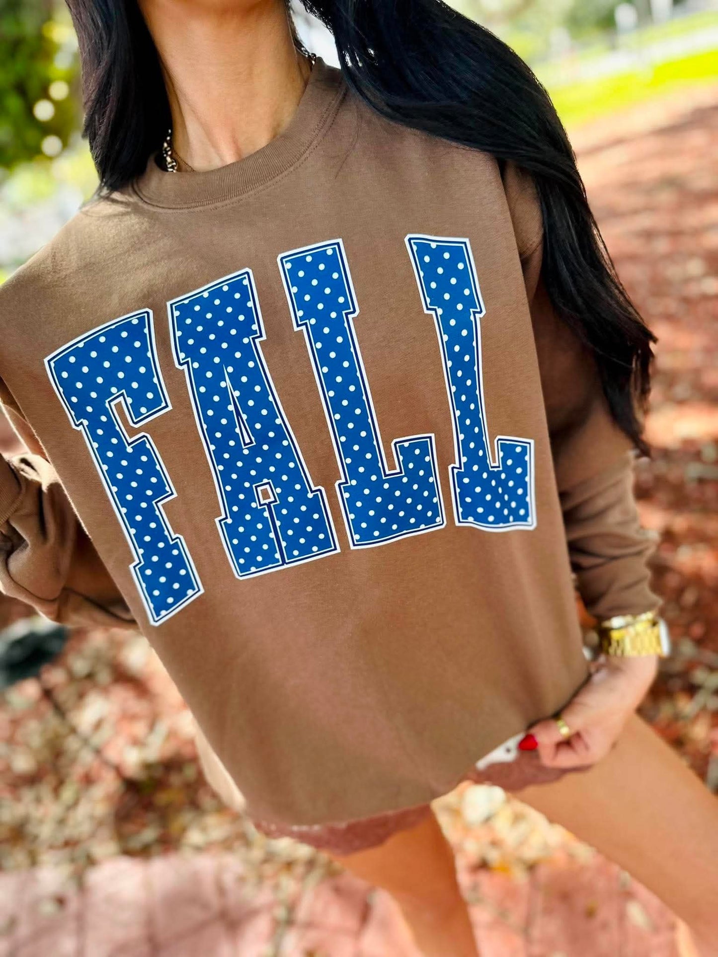 A woman wearing a brown crewneck sweatshirt with the word 'FALL' printed in large blue polka-dot varsity letters outlined in white, set against an outdoor brick path background.