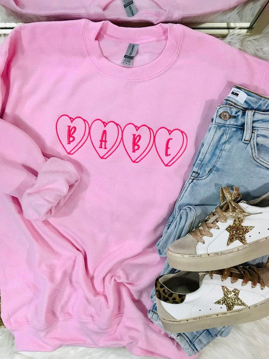 A pink embroidered sweatshirt with the word 'BABE' in heart shape on the front, displayed flat with a pair of blue jeans and white sneakers on top.