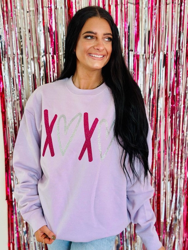 A woman smiling, wearing a lavender multicolor glitter vinyl sweatshirt with a heart and 'XOXO' text on it, standing in front of a pink glittery background.