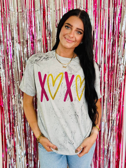 A person wearing a gray t-shirt with hot pink and gold glitter hearts design, standing in front of a pink glittery background.